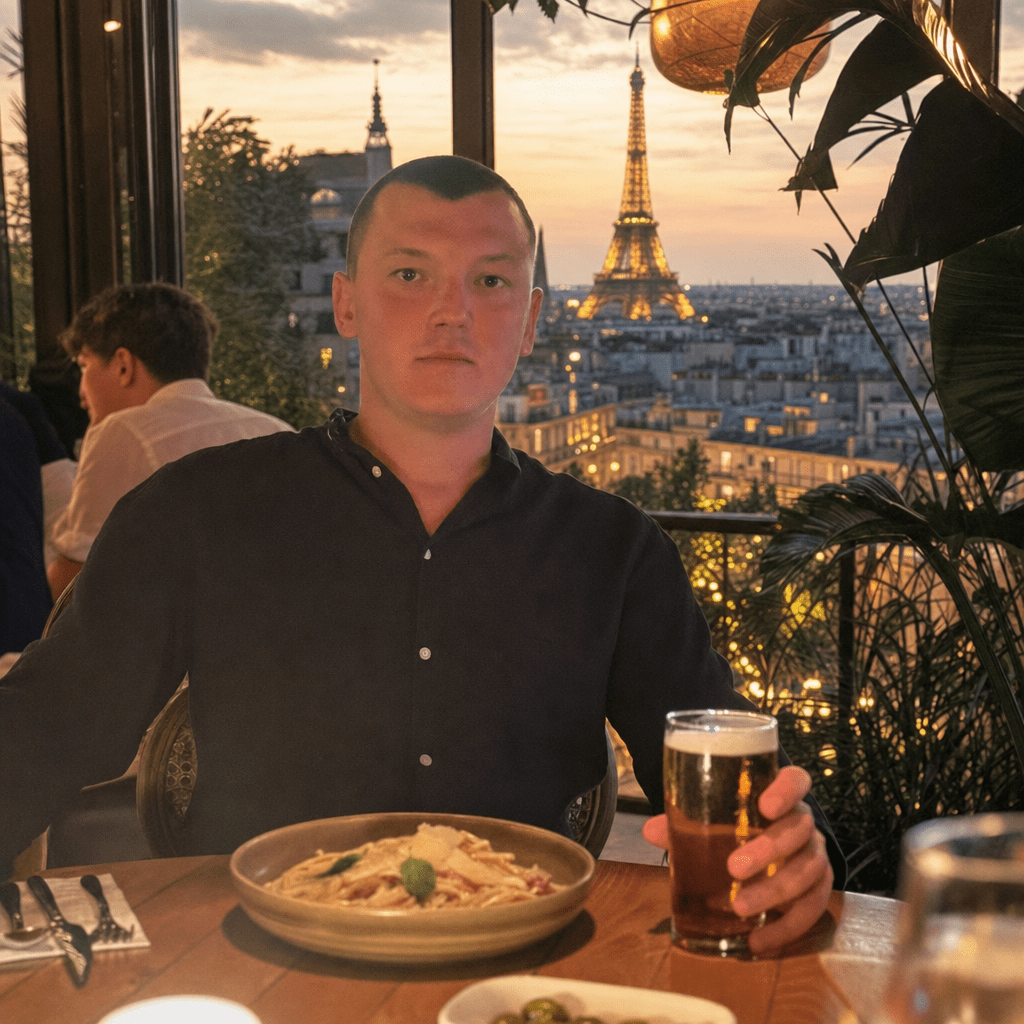 Photorealistic edit of the provided image: keep the person exactly the same—pose, facial features, clothing (dark button-up shirt), and all foreground elements (table setting, plate of pasta, glass with drink, chair/umbrella elements, and any visible plants) with the same natural shadows and warm golden-hour restaurant lighting. Replace only the background/window view behind the person with an upscale Paris evening scene: floor-to-ceiling restaurant windows or open terrace framing a Paris cityscape. The Eiffel Tower must be clearly visible in the distance as the focal point, with realistic scale and perspective matching the original camera angle. Add subtle warm golden-hour haze over the city, soft atmospheric depth, and realistic window reflections/glass glare consistent with the foreground lighting direction. Ensure natural, consistent lighting on the subject and minimal artifacts around the subject’s edges and hands. Overall look should be indistinguishable from a real photograph.